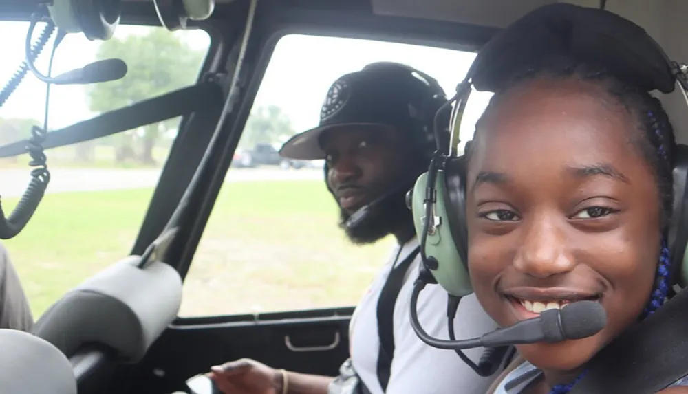 A smiling young person wearing a headset is seated in a vehicle with another person wearing a headset visible in the background