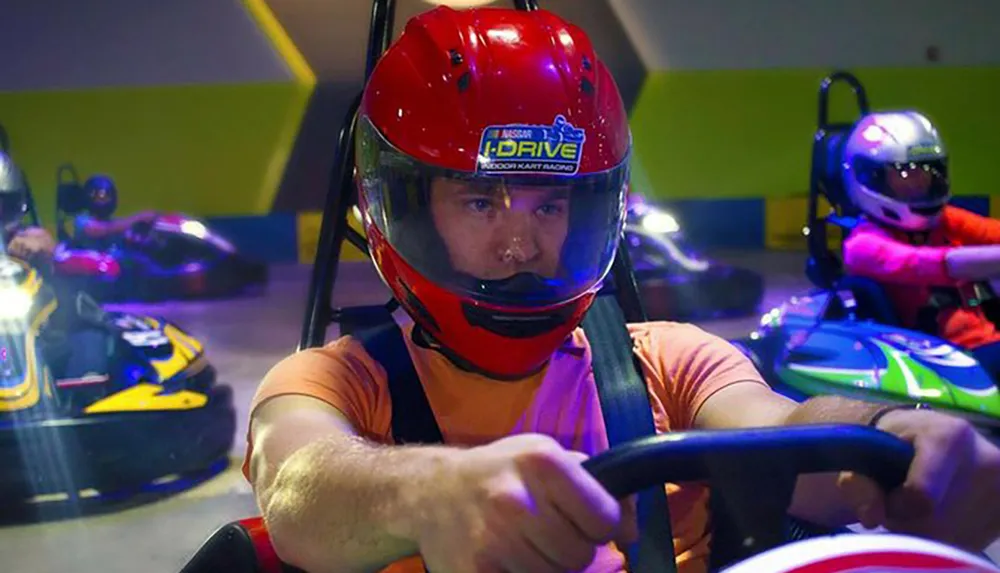 A person wearing a red helmet is driving a go-kart on an indoor track with other racers visible in the background
