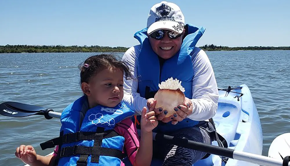 A child and an adult wearing life jackets are on a kayak with the adult showing a sea creature to the child