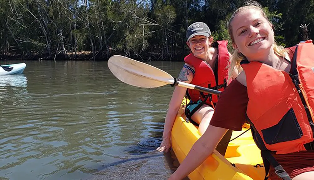 Two people are smiling and posing for the camera while kayaking on a river wearing red life vests and holding paddles