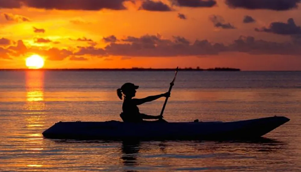 A person kayaks in tranquil waters against the backdrop of a vibrant sunset