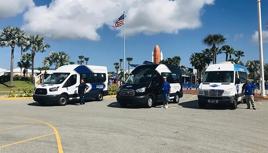 Three shuttle buses are parked with drivers standing by them in a sunny parking lot, with palms and a space shuttle display in the background under a clear blue sky.