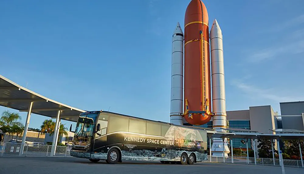 A Kennedy Space Center branded bus is parked in front of a towering display of a Space Shuttles orange external fuel tank and two solid rocket boosters