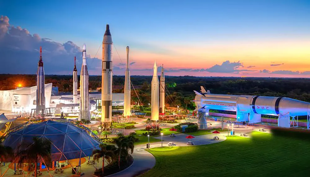 The image depicts an outdoor exhibition of various historical rockets and a space shuttle at a space-themed museum during twilight
