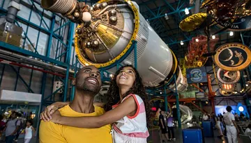 A smiling man and a young girl look up in awe at a spacecraft exhibit in a space museum.