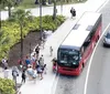 A group of passengers is boarding a red city bus at a stop with cars passing by on the adjacent lane