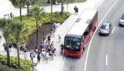 A group of passengers is boarding a red city bus at a stop, with cars passing by on the adjacent lane.