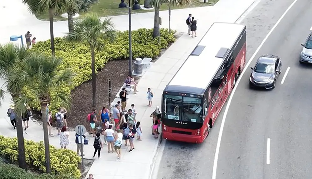 A group of passengers is boarding a red city bus at a stop with cars passing by on the adjacent lane