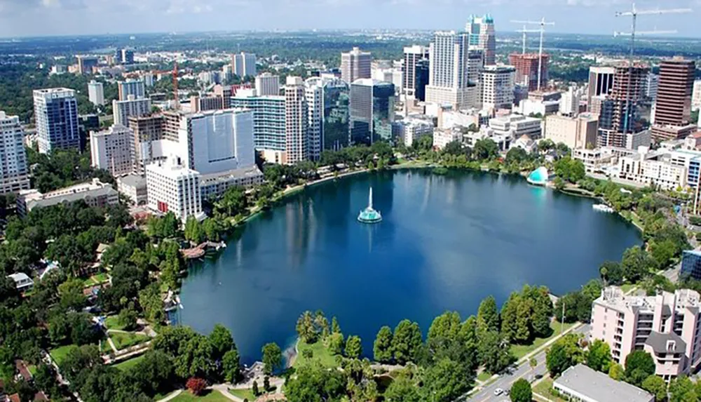 This image is an aerial view of a downtown skyline with modern buildings surrounding a serene lake which features a fountain in its center