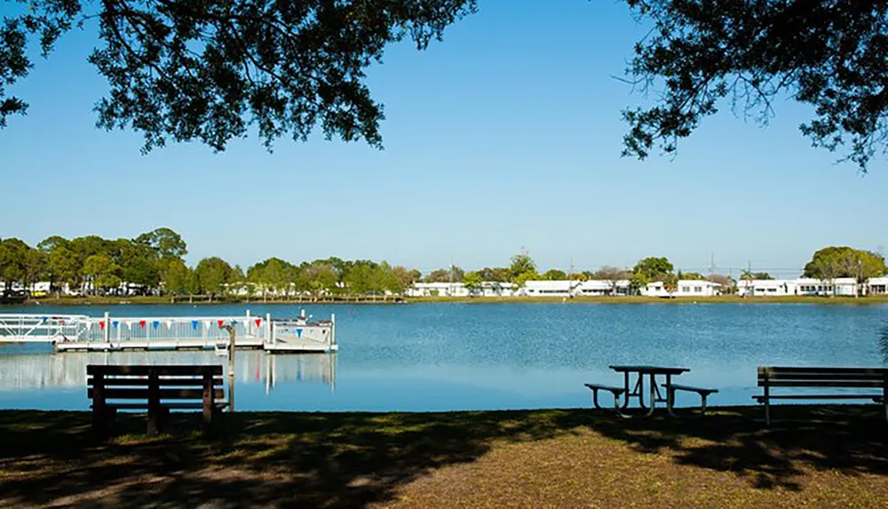 The image shows a serene lakeside scene with a dock pedal boats a wooden bench a picnic table under trees and a clear blue sky