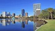 The image depicts a scenic view of a modern city skyline reflected in a tranquil body of water, framed by lush greenery and a clear blue sky.
