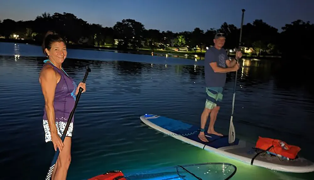 Two people are paddleboarding on a calm body of water at dusk with one of the boards featuring neon underglow lighting