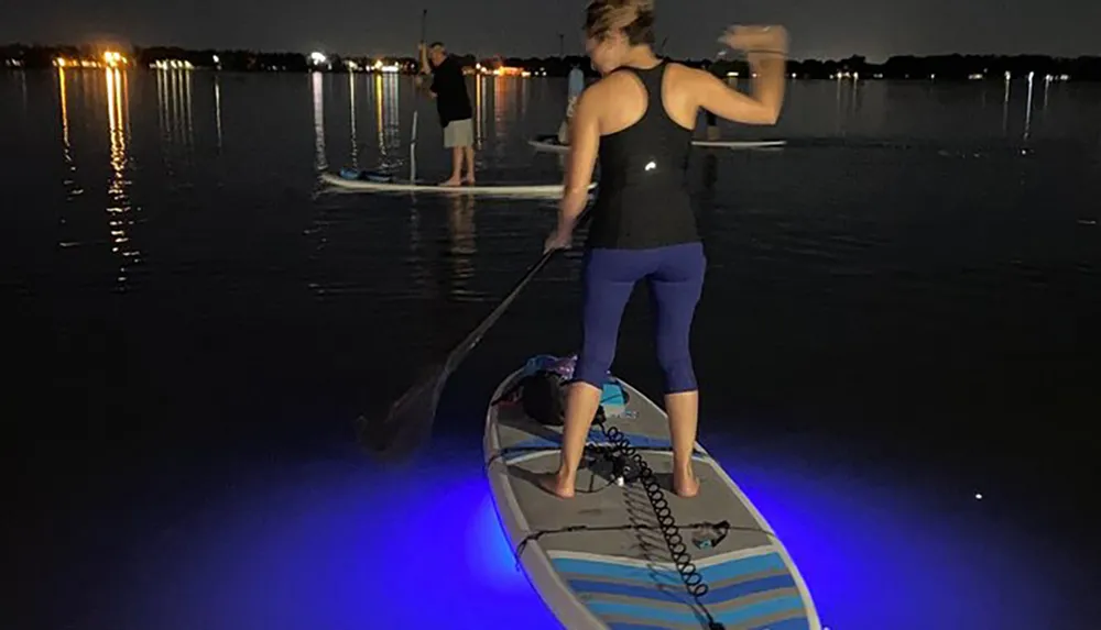 Two people are paddleboarding at night with one board illuminated by a blue light giving a serene glow on the water