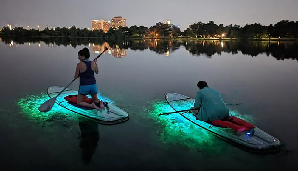 Two people are paddleboarding at night on a calm body of water illuminated by lights attached to their boards with a city skyline in the background