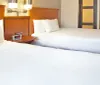 The image shows a tidy hotel room featuring two double beds with white linens a nightstand with a digital alarm clock and a glimpse of the rooms entrance area