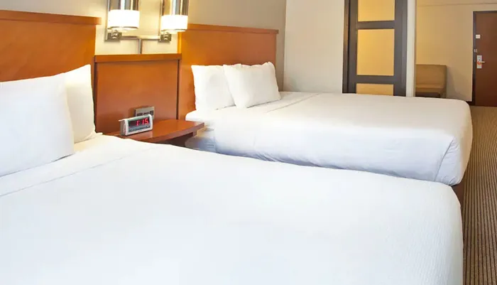 The image shows a tidy hotel room featuring two double beds with white linens a nightstand with a digital alarm clock and a glimpse of the rooms entrance area