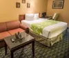 The image shows a hotel room with two twin beds covered with white and green linens flanked by bedside tables and lamps with a framed picture on the wall and a small dining table with chairs to one side