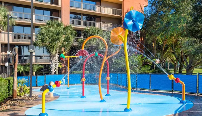 A colorful childrens splash pad with various water features is situated in front of a multi-story building with balconies
