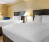 The image shows a neatly made king-sized bed with white linens in a well-lit hotel room flanked by two nightstands with lamps and a blue lounge chair in the corner