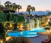 The image depicts a luxurious resort-style swimming pool with artificial waterfalls surrounded by palm trees and lush vegetation captured during twilight