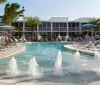 The image shows a tranquil hotel pool area with fountains surrounded by palm trees and loungers under a clear sky
