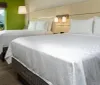 The image shows a bright and neatly arranged hotel room with two large beds a green accent wall modern lighting and a view of the outside from a large window