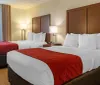The image shows a neatly arranged hotel room with two double beds featuring white linens and red decorative bed runners flanked by nightstands and lamps with a window in the background