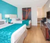 This image showcases a neatly arranged hotel room with two beds featuring white and turquoise bedding a large turquoise accent wall a desk with an orange lamp a mounted television and a decorative piece of wall art