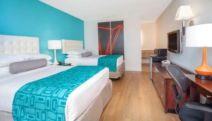 This image showcases a neatly arranged hotel room with two beds featuring white and turquoise bedding a large turquoise accent wall a desk with an orange lamp a mounted television and a decorative piece of wall art