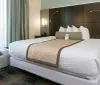 A modern hotel room is furnished with a large bed clean linens flanked by wall-mounted lamps and complemented with a long cushioned headboard and a decorative bed runner creating a welcoming atmosphere
