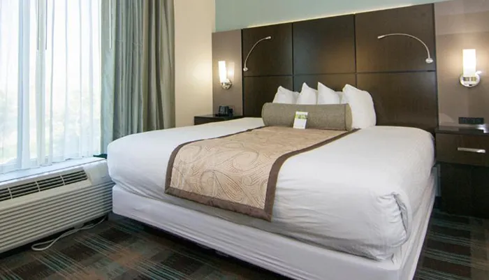 A modern hotel room is furnished with a large bed clean linens flanked by wall-mounted lamps and complemented with a long cushioned headboard and a decorative bed runner creating a welcoming atmosphere