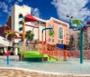 This image shows a vibrant and colorful childrens water play area with slides and splash buckets in front of a hotel building under a clear blue sky