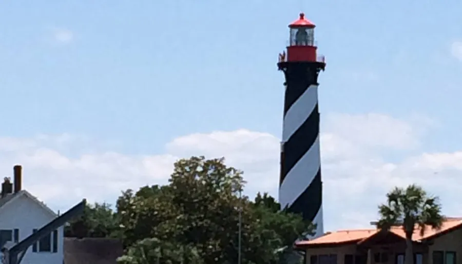 A tall, spirally-striped black and white lighthouse with a red top stands prominently under a blue sky with scattered clouds, with houses and trees in the foreground.