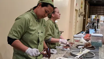 Two people in chef uniforms are working diligently in a kitchen, one spreading chocolate on a dessert.