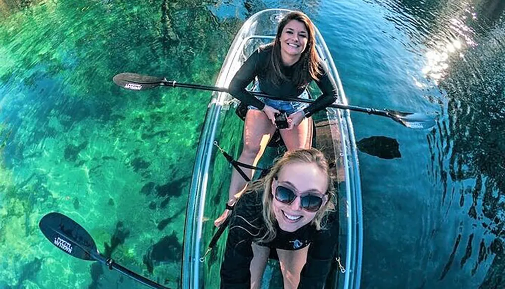 Two women are smiling and enjoying a clear water kayaking experience in a transparent kayak