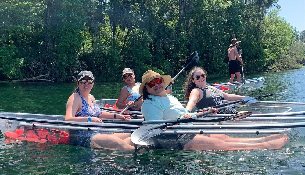 A group of smiling people are enjoying kayaking and paddleboarding on a sunny day in clear water with lush greenery in the background