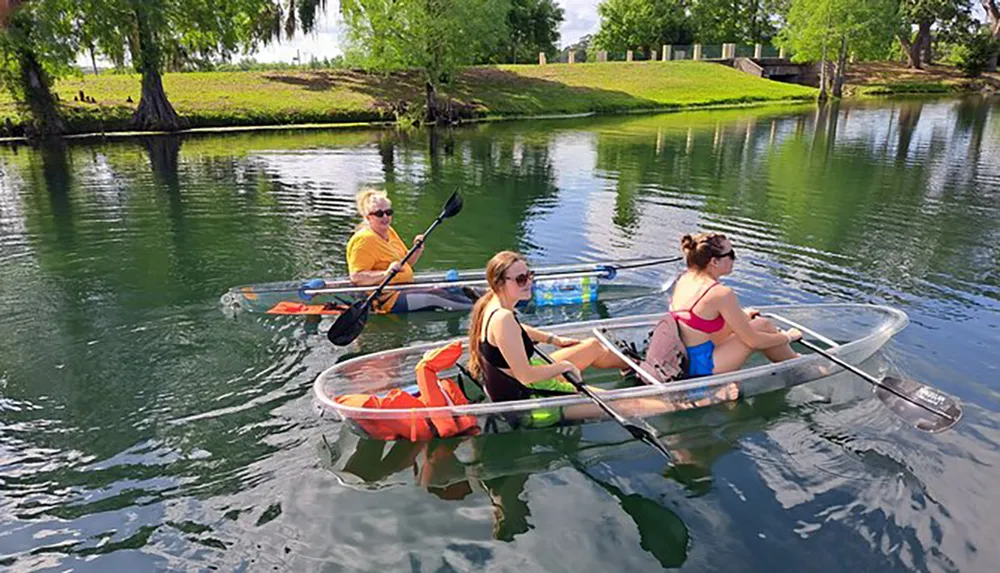 Three people are paddling in transparent kayaks on a calm body of water offering a clear view of the underwater scene