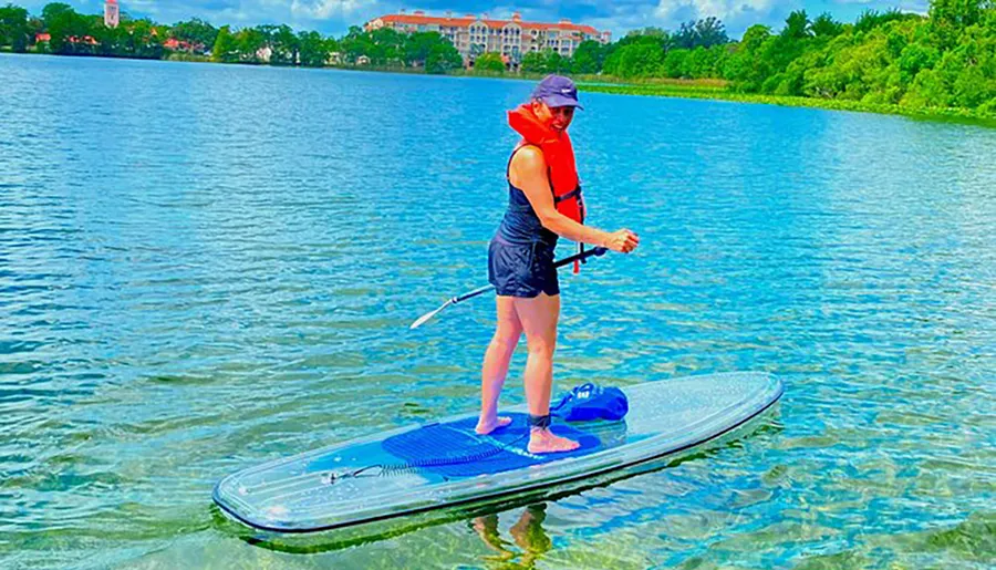 A person wearing a red life vest stands on a paddleboard in a body of water, holding a paddle, with trees and a building in the background.