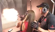 A person is firing a handgun with a large muzzle flash at an indoor shooting range while an instructor stands by her side.