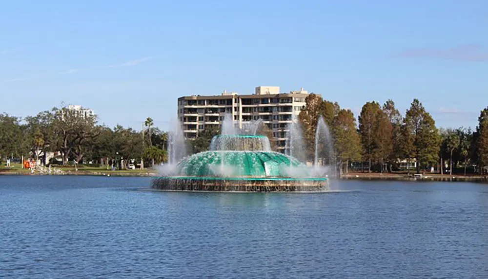 A large tiered fountain with a turquoise-colored lower tier sprays water into the air set against the backdrop of a lake palm trees and buildings under a blue sky
