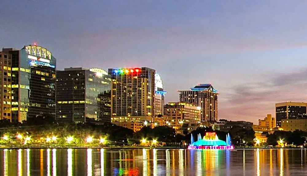The image shows a vibrant city skyline at dusk with illuminated buildings reflecting off the waters surface accented by a colorful lit fountain in the foreground