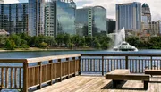 A waterfront view from a wooden deck featuring a city skyline and a fountain in the center of a lake under a blue sky.