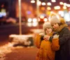 Two children warmly dressed in winter clothing are hugging affectionately on a city street lit by the glow of streetlights and storefronts at dusk or night