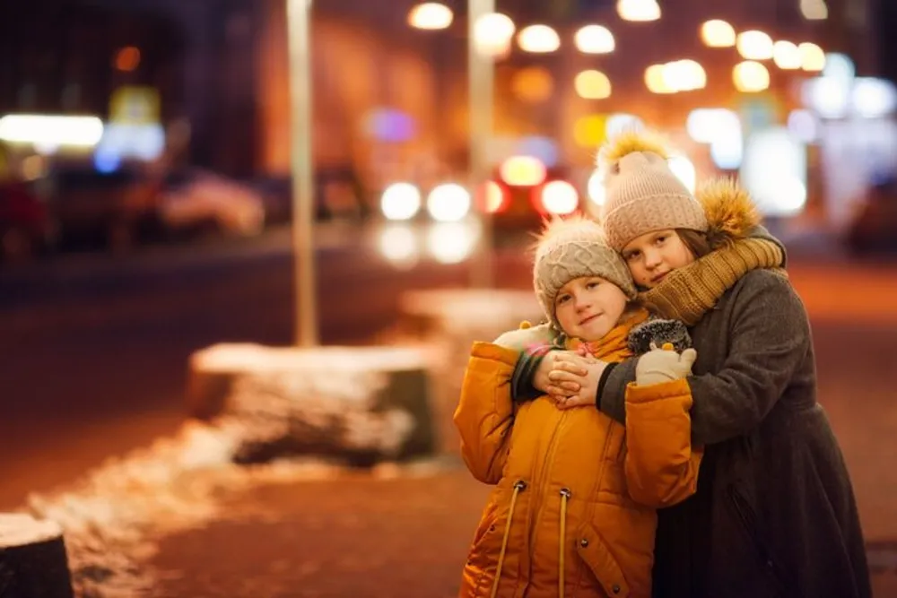 Two children warmly dressed in winter clothing are hugging affectionately on a city street lit by the glow of streetlights and storefronts at dusk or night