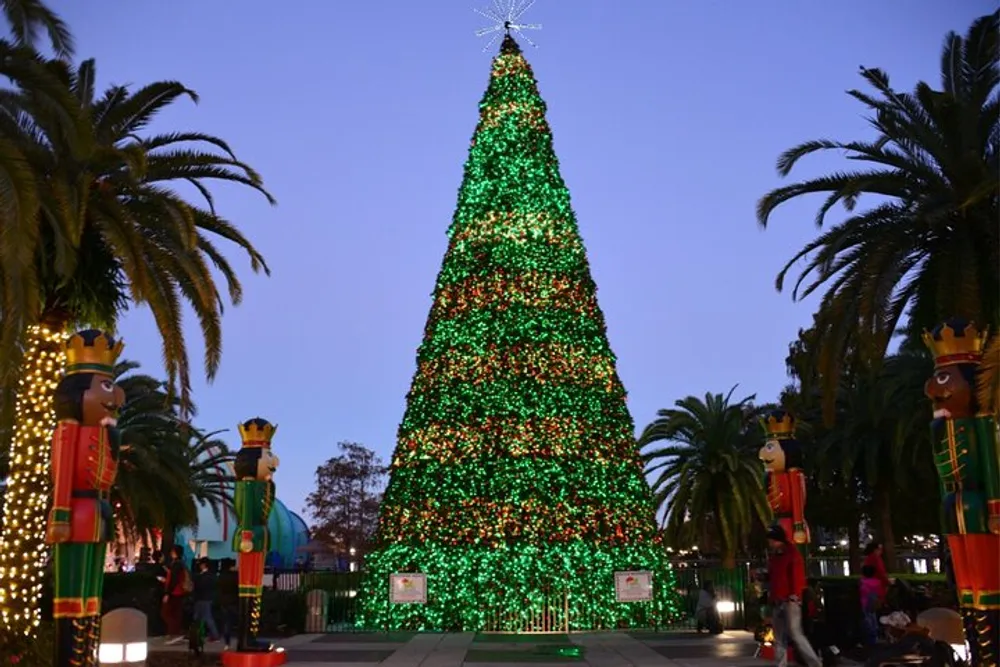 A large illuminated Christmas tree is adorned with red and green lights flanked by two nutcracker statues against a backdrop of twilight and palm trees