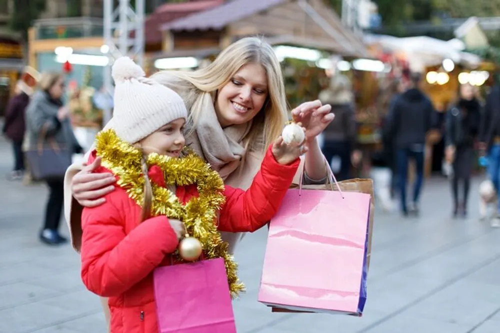 A smiling woman and a young girl who is holding a golden tinsel garland and a shopping bag appear to be enjoying a festive outdoor holiday market