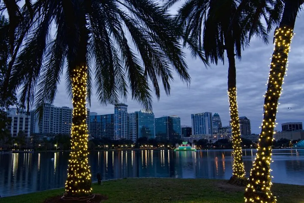 Palm trees wrapped in twinkling lights frame a view of a serene lake with a cityscape backdrop under a dusky sky