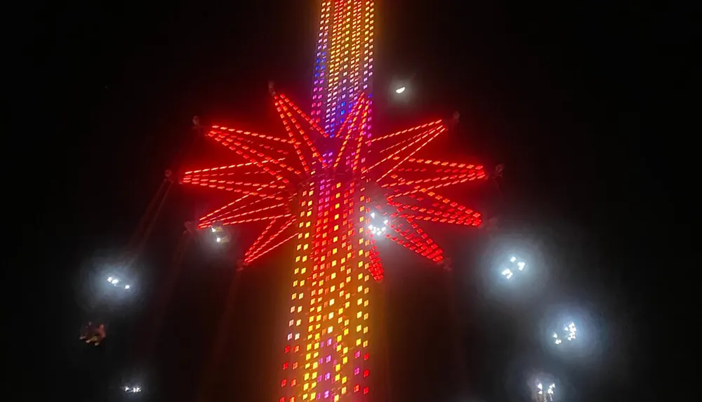 The image captures a brightly illuminated vertical amusement ride at night adorned with vibrant red and blue lights and surrounded by a few conventional spotlights