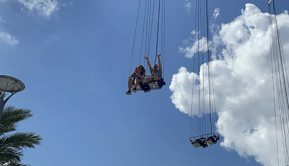 Two people are swinging high up in the air on a swing ride on a sunny day with clear blue skies and fluffy clouds above