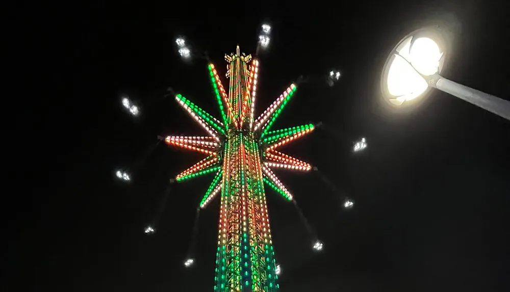 The image shows an illuminated festive tower adorned with green and red lights standing out against the dark night sky accompanied by a bright streetlamp to the right